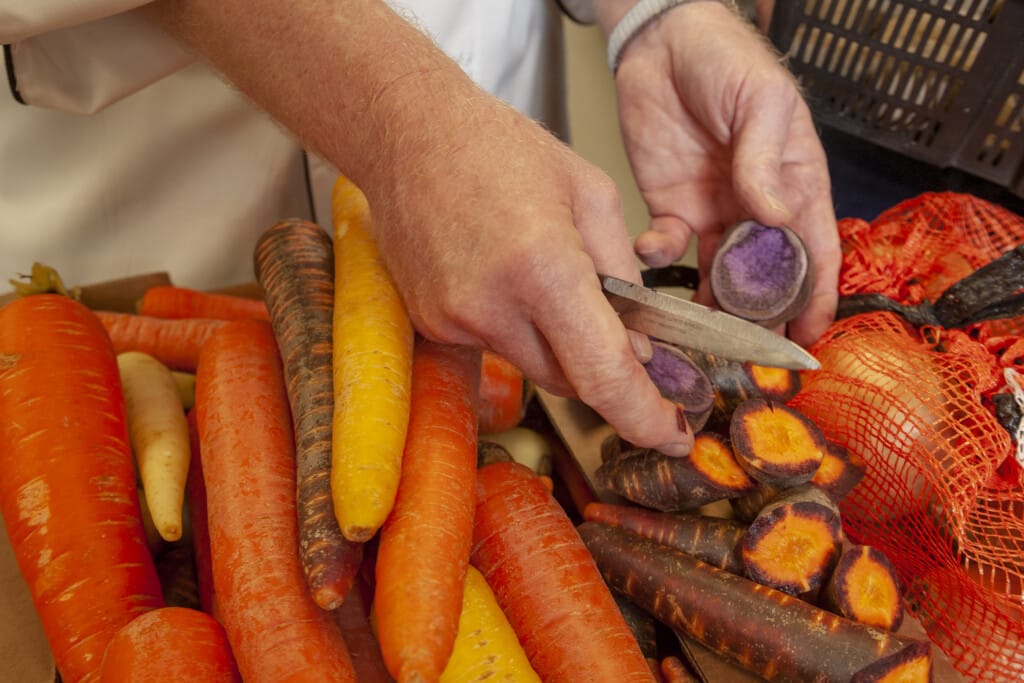 Purple potatoes and yellow, purple and orange carrots being cut by a cook in a restaurant kitchen.
